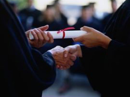   At graduation, two people in grad gowns are standing. They are shaking hands and receiving diploma